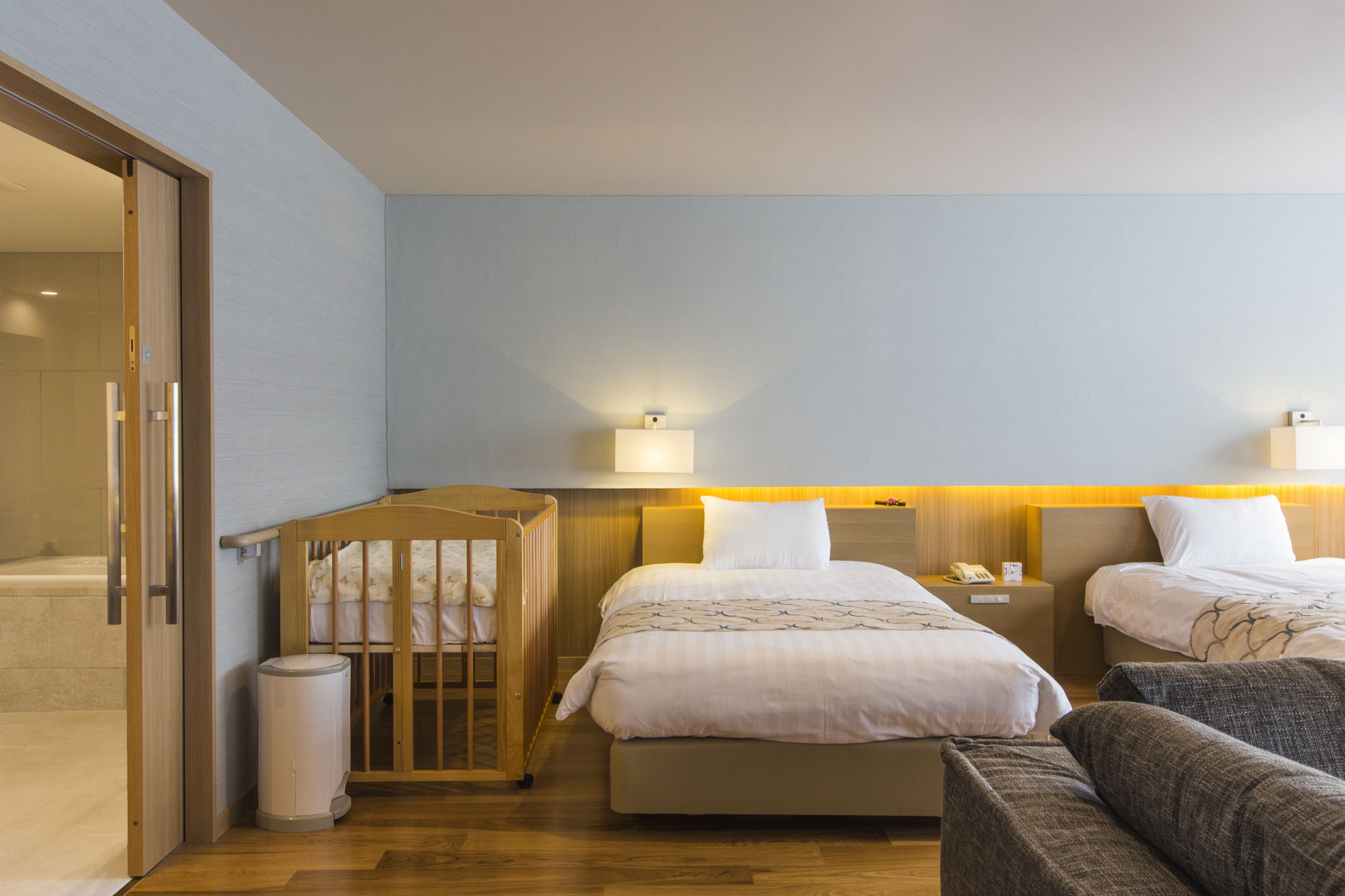 Habitación de hotel con dos camas, una cuna de madera, un cubo de basura y una mesita con lámpara.
