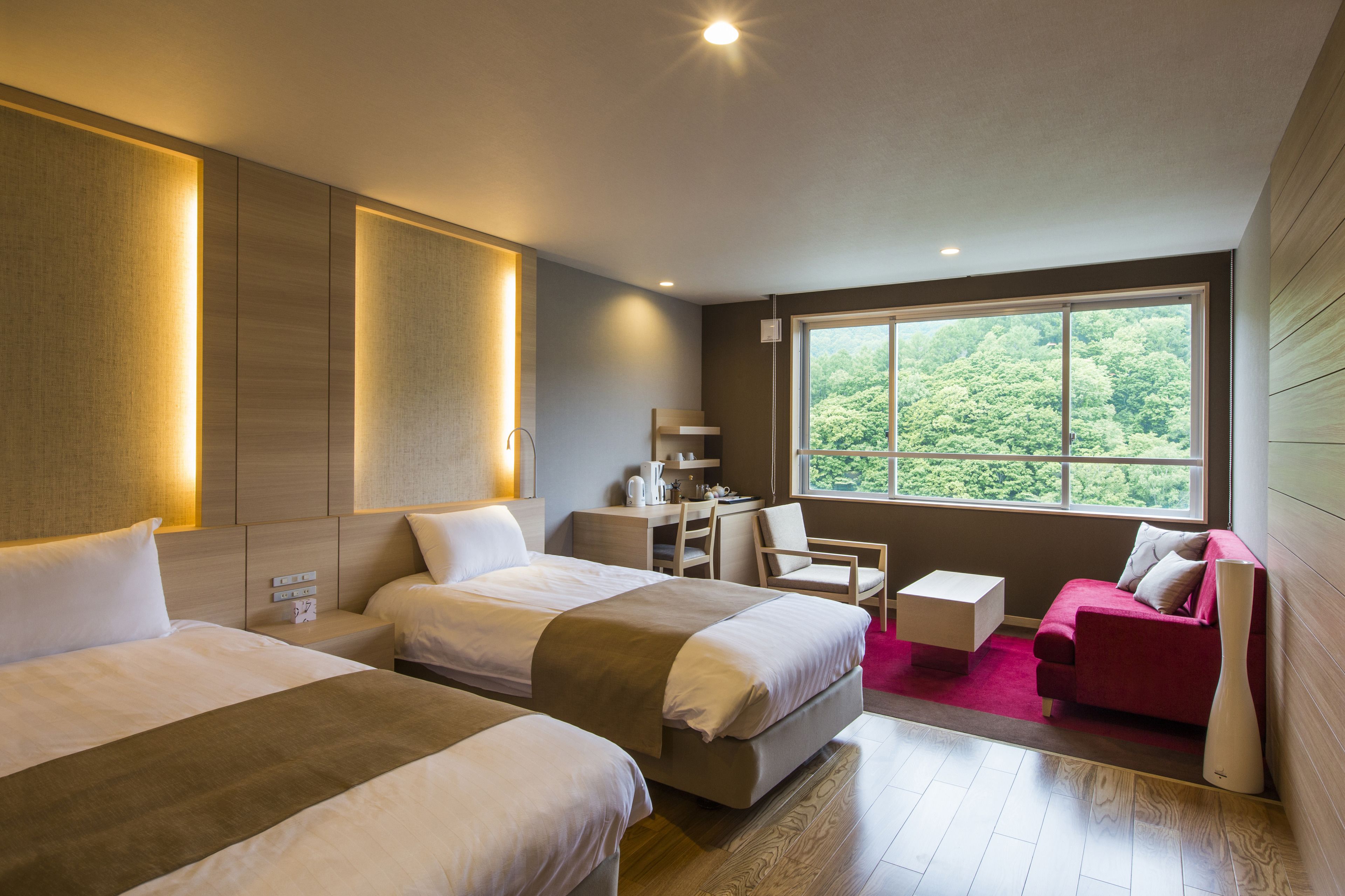 A hotel room with two beds, a desk, a chair, a sofa, and a large window with a view of greenery.