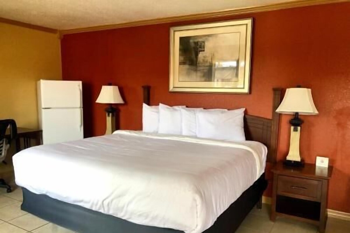 Une chambre d’hôtel comprenant un lit, deux tables de chevet, une commode, une lampe, un tableau accroché au mur et un réfrigérateur.