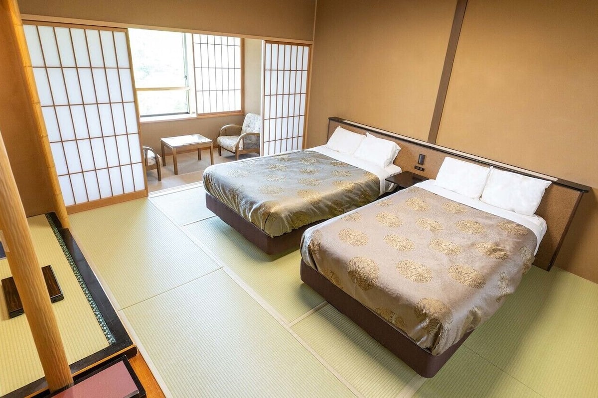 A room with tatami flooring, two beds with patterned bedding, a chair, and a sliding door.