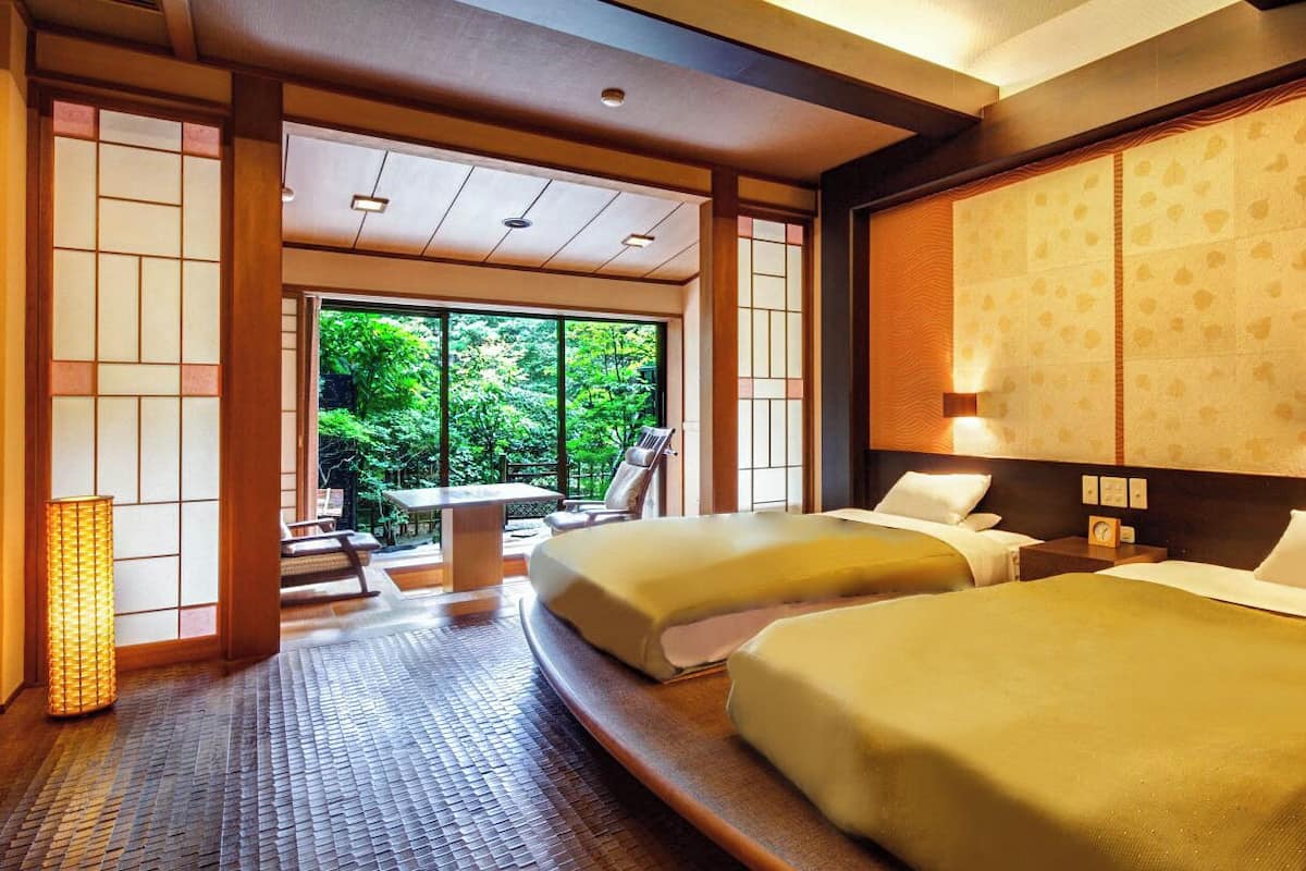 A traditional Japanese-style hotel room with tatami flooring, two beds, a sliding door, and a view of greenery outside.