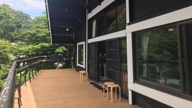 A wooden deck with a railing, large windows, and a view of greenery.
