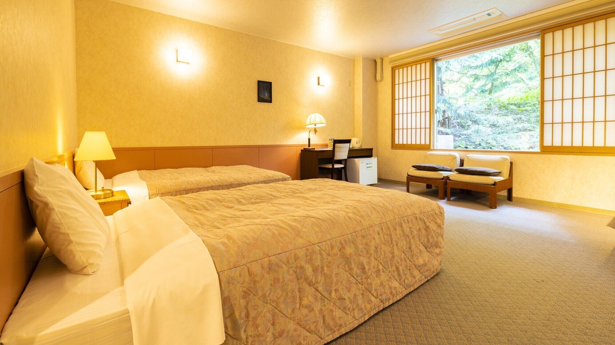 Ein Hotelzimmer mit zwei Betten, einem Schreibtisch, einem Stuhl und einem Fenster mit Blick auf Grünflächen.