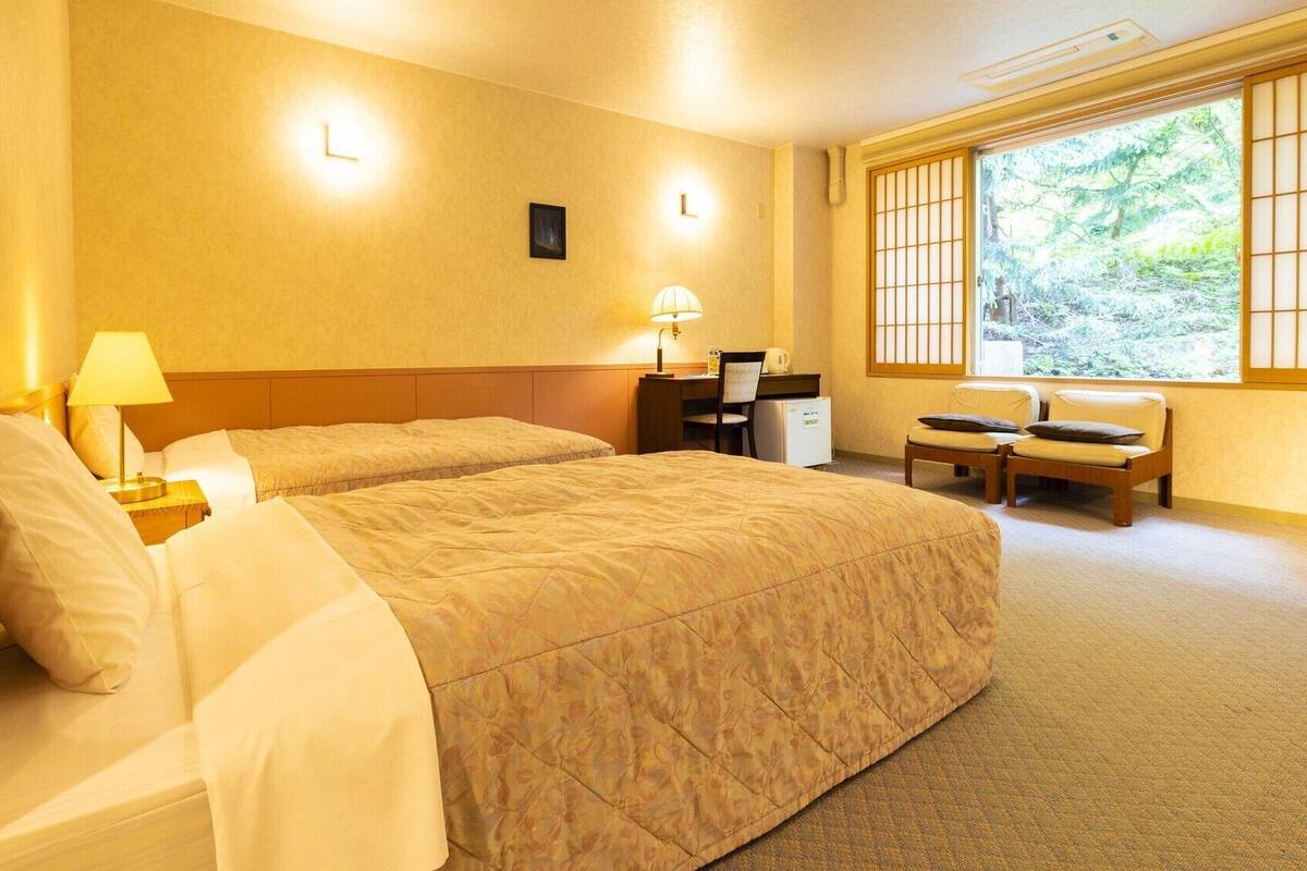 A hotel room with two beds, a desk, a chair, and a window with a view of greenery.