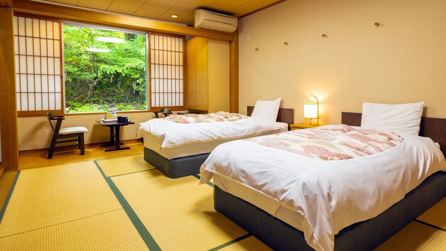 Ein Hotelzimmer mit zwei Betten, einem kleinen Tisch, einem Stuhl und Blick auf Grün hinter dem Fenster.