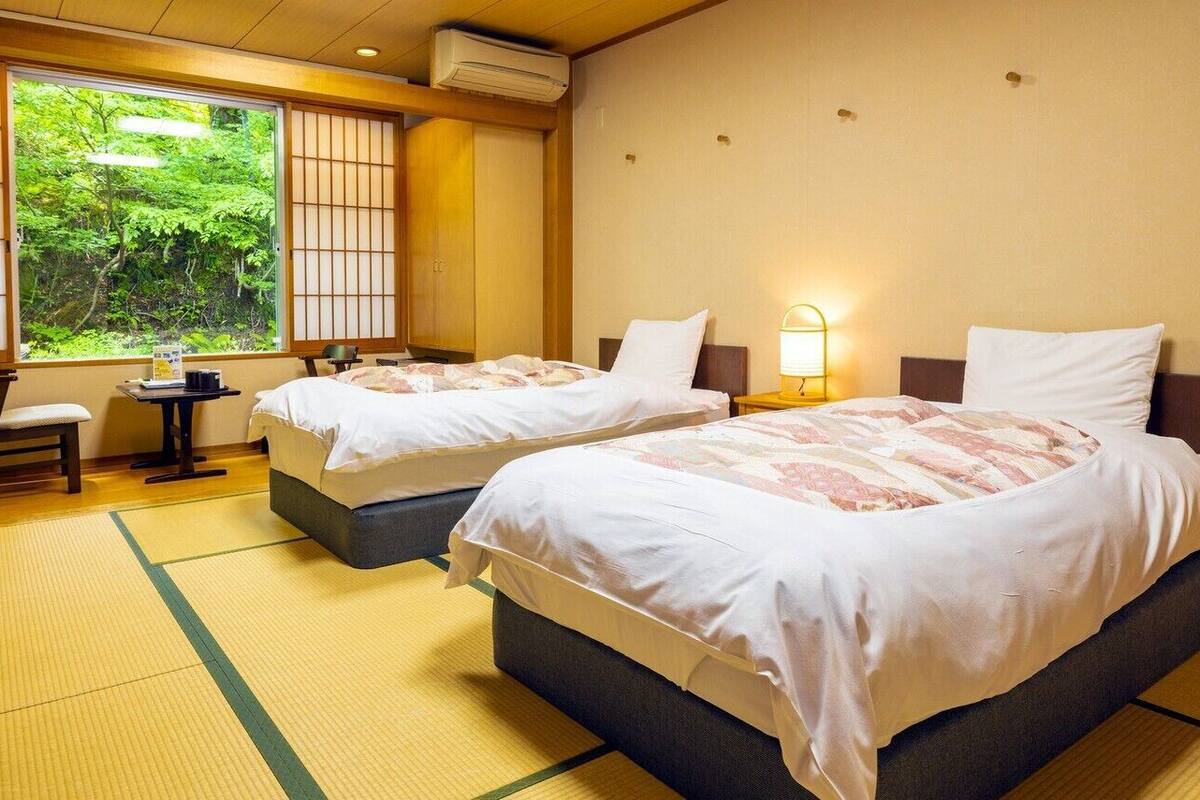 A hotel room with two beds, a small table, a chair, and a view of greenery through the window.