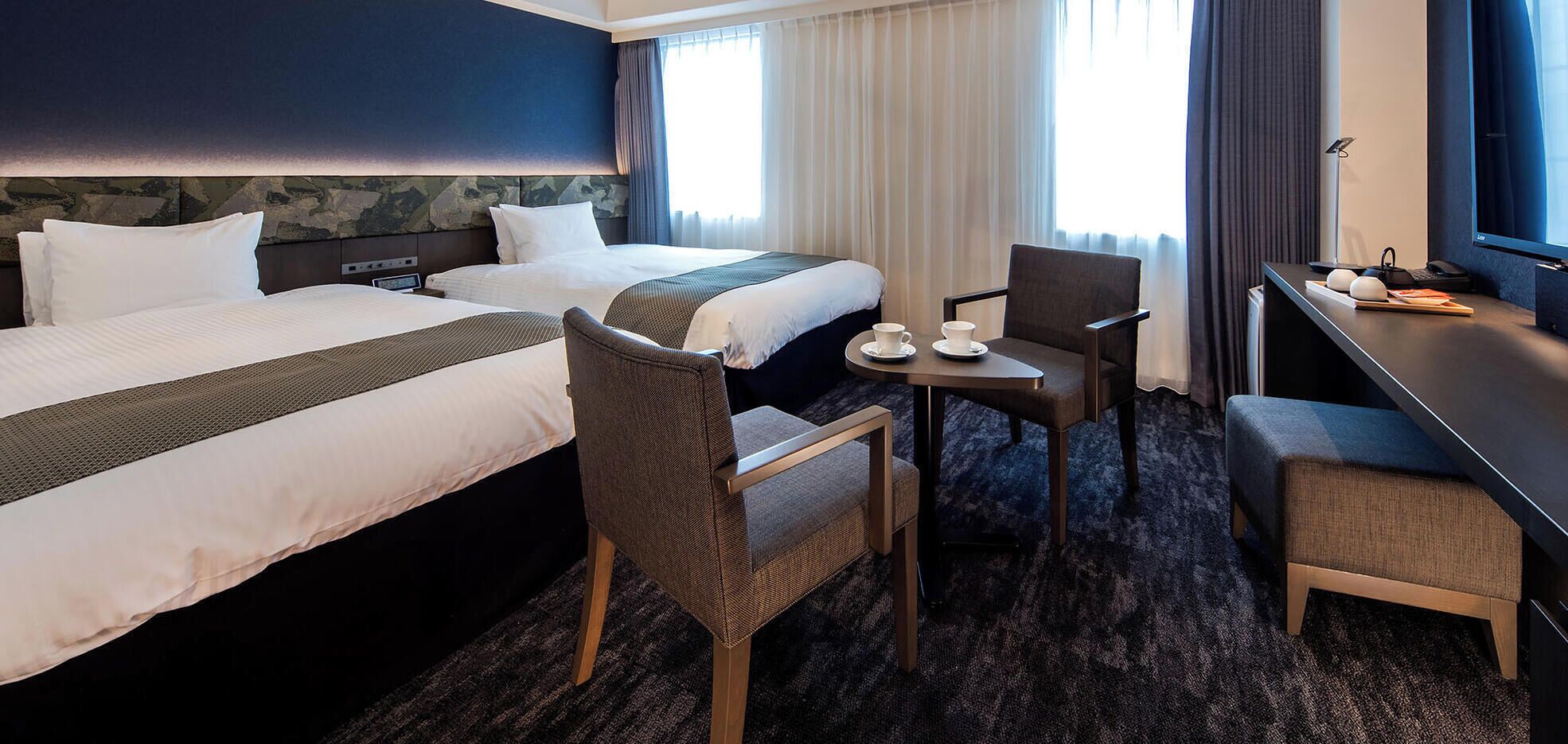 Et hotelværelse med to senge, et lille bord med kop og underkop, en stol og et skrivebord med en computer.