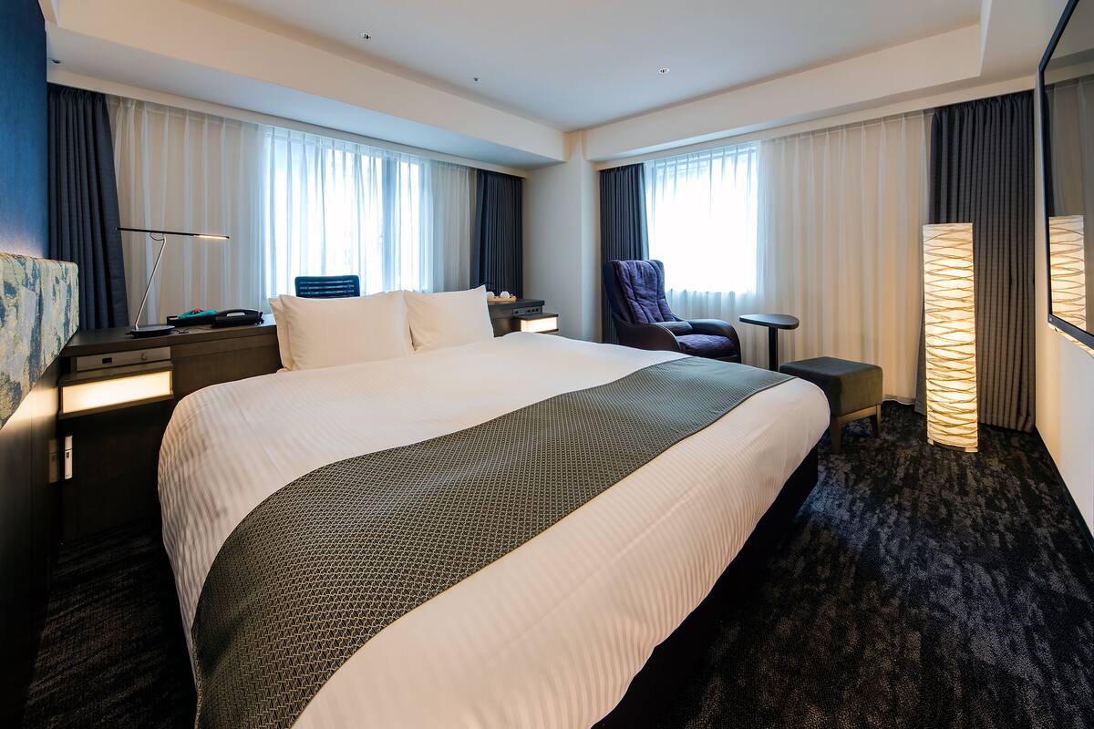 Ein Hotelzimmer mit einem großen Bett, einem Schreibtisch, einem Stuhl, einer Lampe und einem Fenster mit Vorhängen.