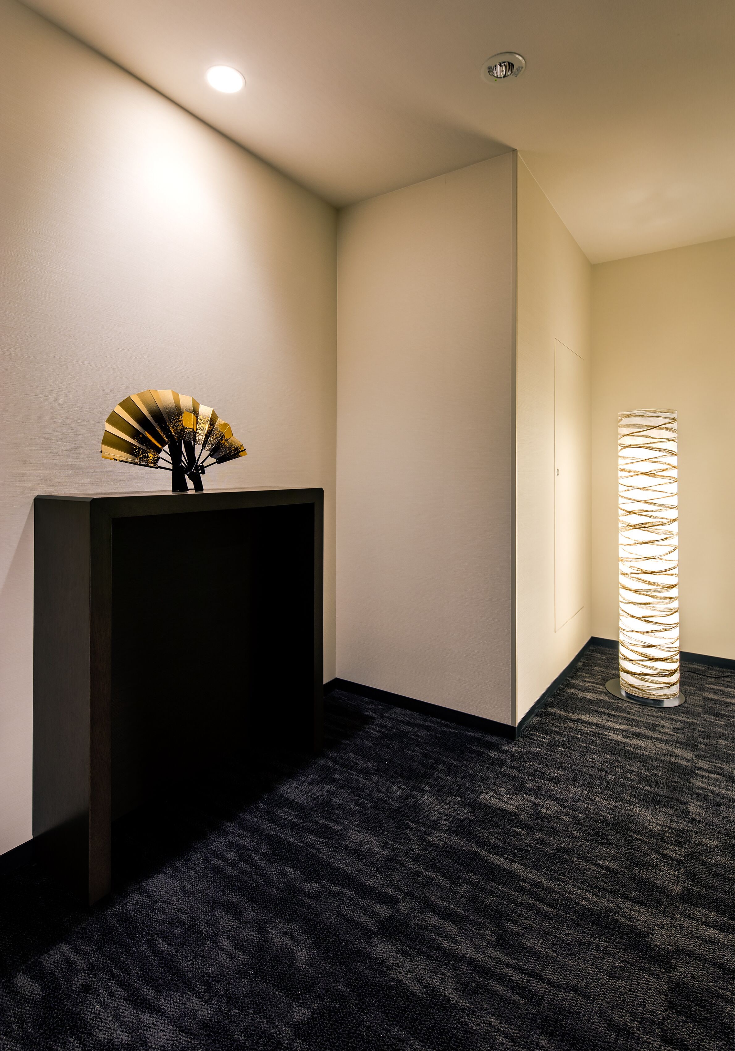 Un vestíbulo de hotel con alfombra oscura, un mostrador de recepción, un abanico decorativo y una columna alta e iluminada.