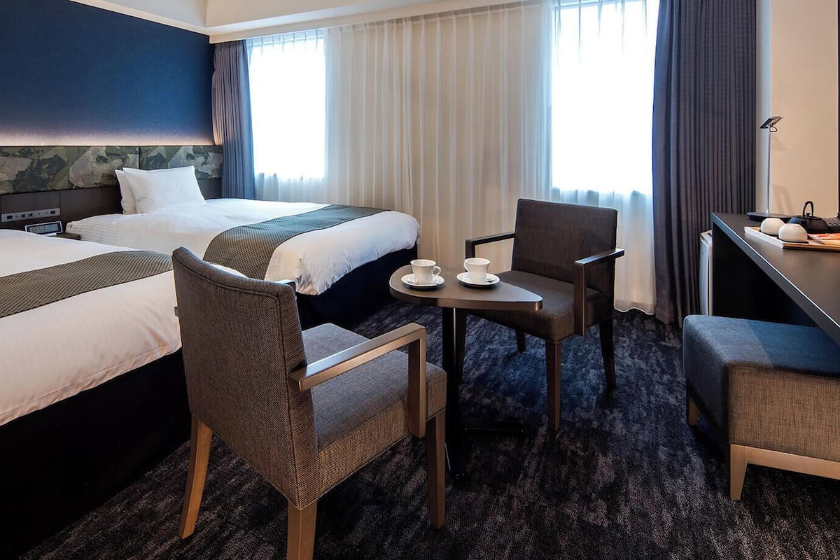Ein Hotelzimmer mit zwei Betten, einem Schreibtisch, einem Stuhl und einem kleinen Tisch mit Tasse und Untertasse.