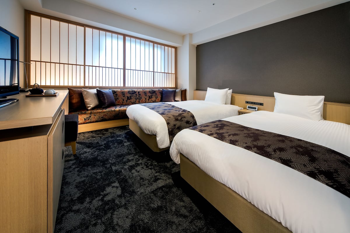 Ein Hotelzimmer mit zwei Betten, einem Schreibtisch, einem Fernseher und einem großen Fenster mit Jalousien.