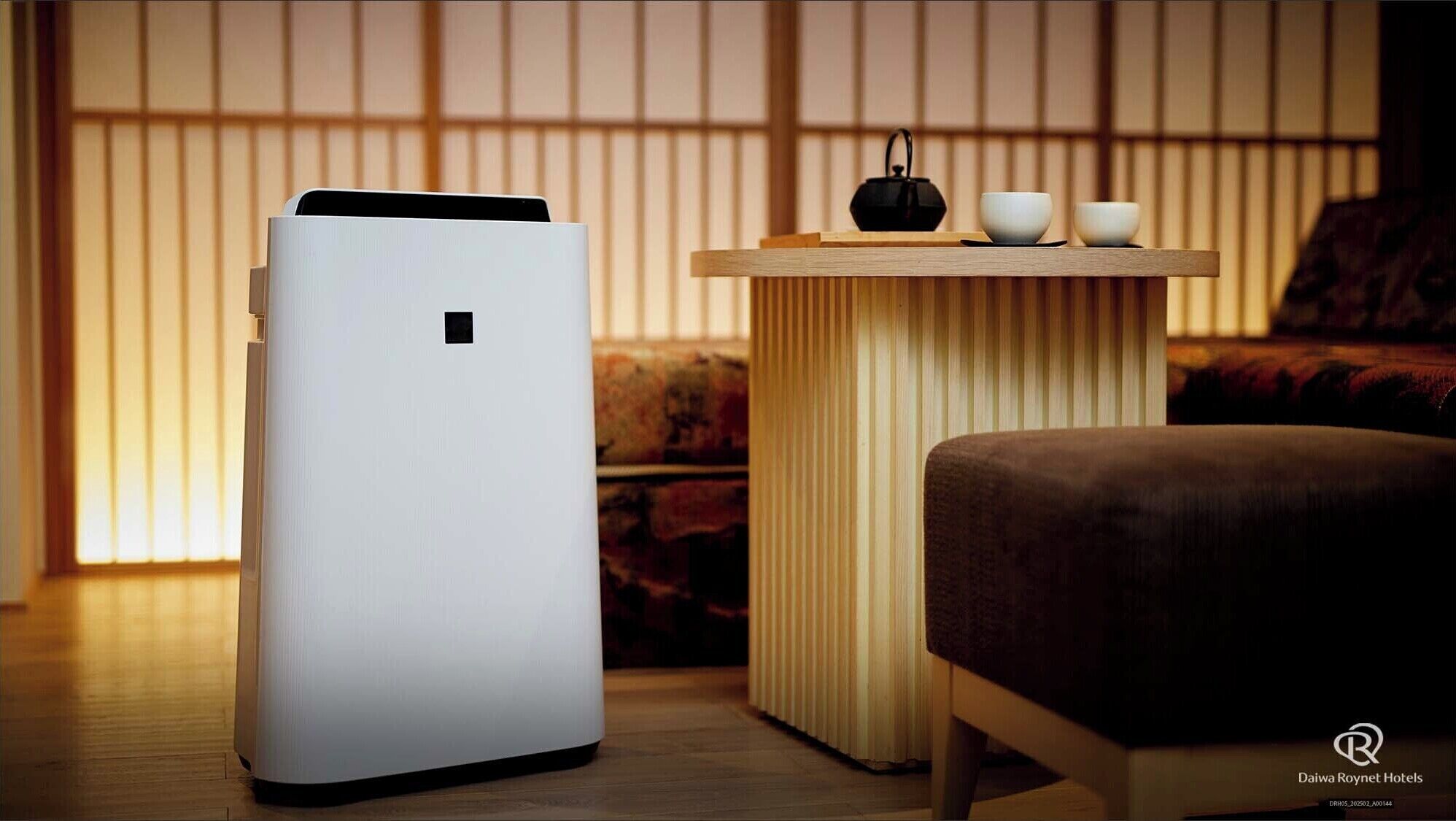 Una habitación moderna con un purificador de aire blanco, una pequeña mesa de madera con una tetera negra, dos cuencos blancos y un sofá marrón.