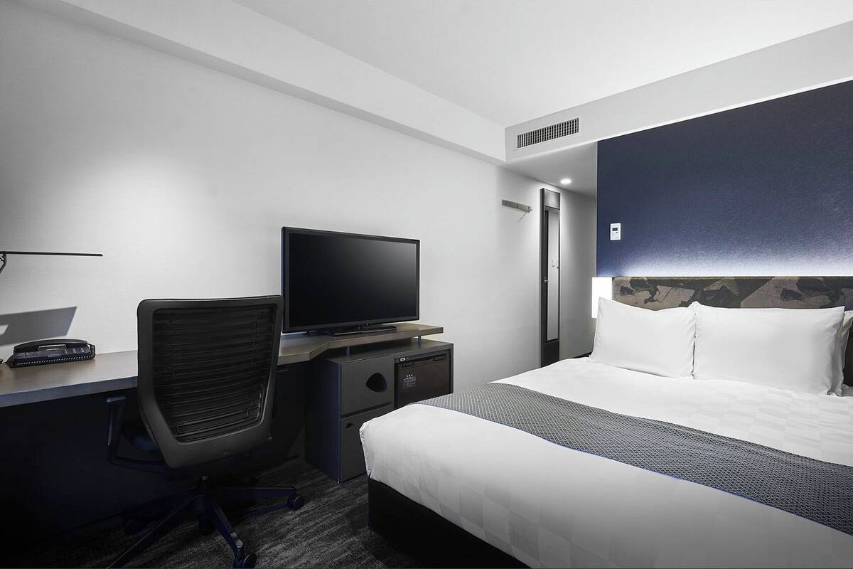 Una habitación de hotel con cama, escritorio, computadora, televisor y una silla.