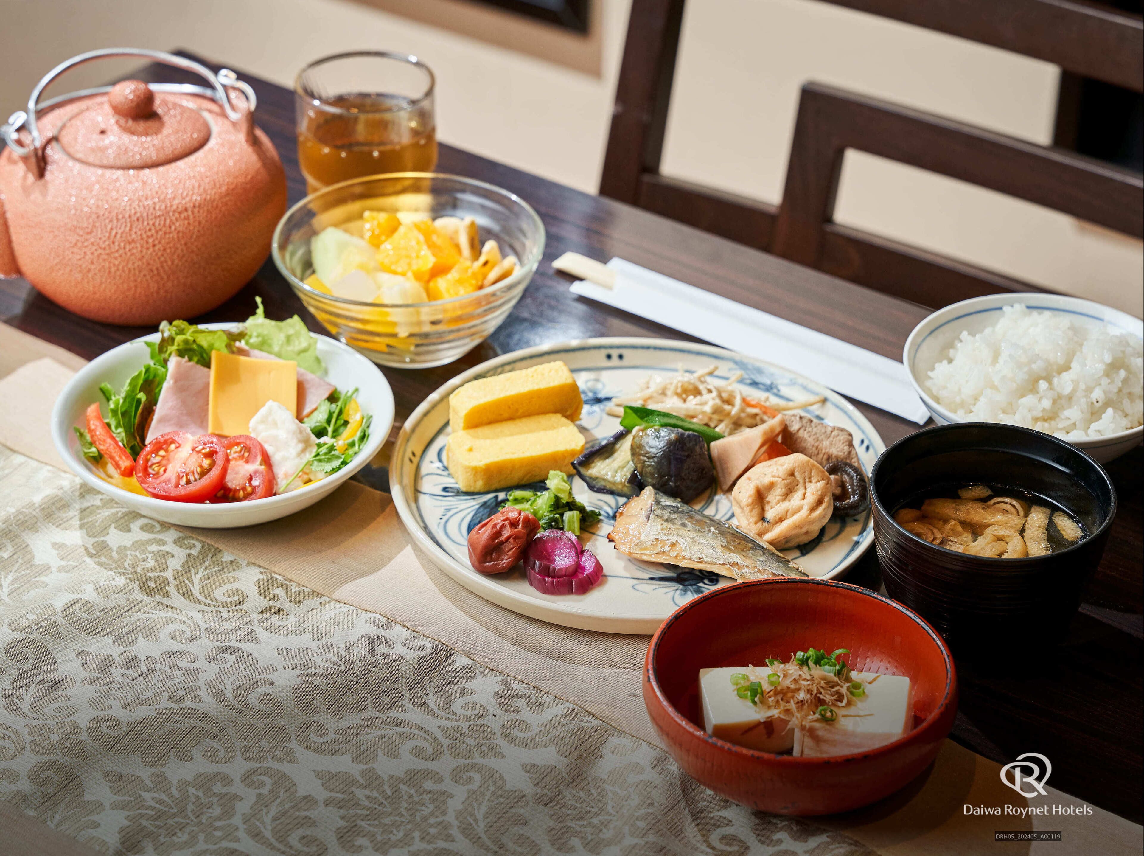 A table with various dishes, including a bowl of rice, a plate with fish and vegetables, a bowl of tofu, and a teapot.