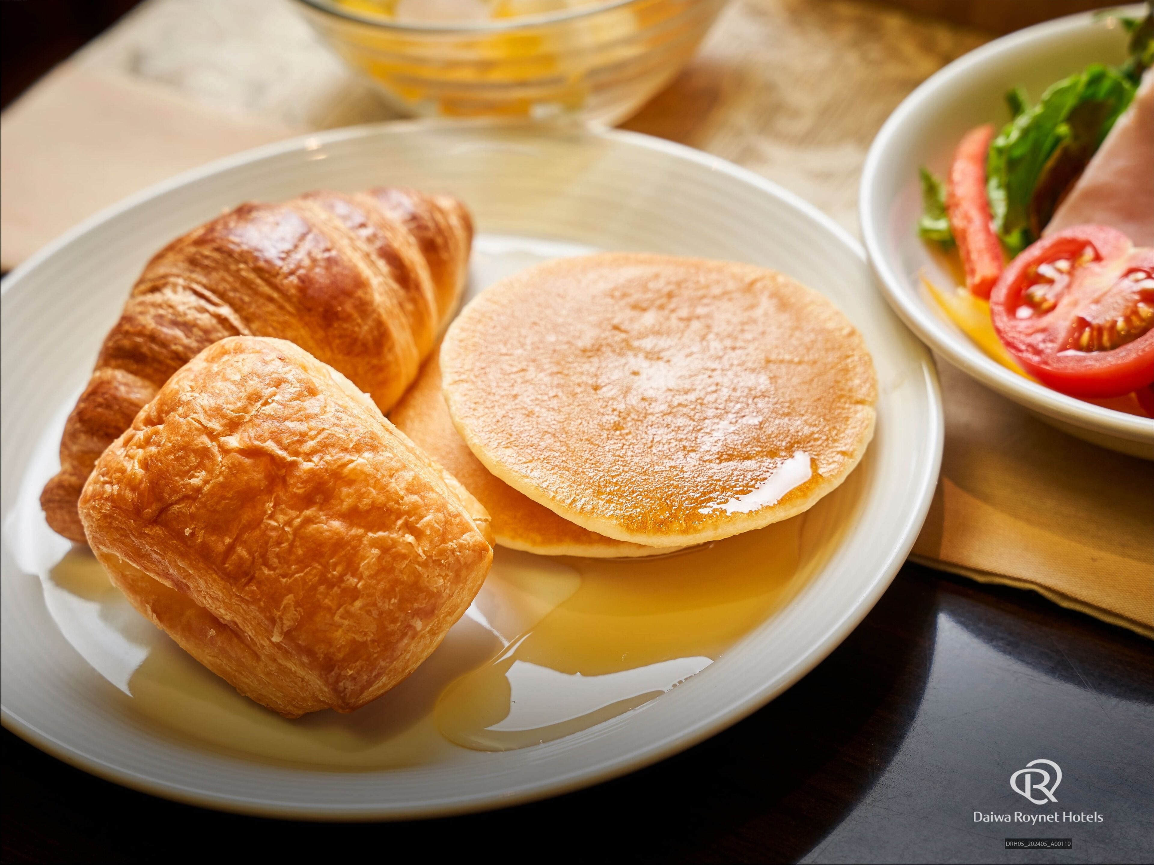 A plate with croissants and pancakes, a bowl of sliced tomatoes, and a glass of orange juice.