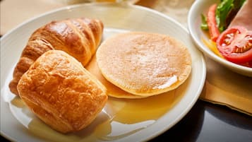 A plate with croissants and pancakes, a bowl of sliced tomatoes, and a glass of orange juice.