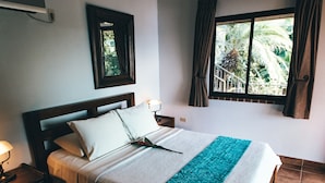 Un dormitorio con cama, mesita de noche, ventana con cortinas, aire acondicionado instalado en la pared y un espejo enmarcado.