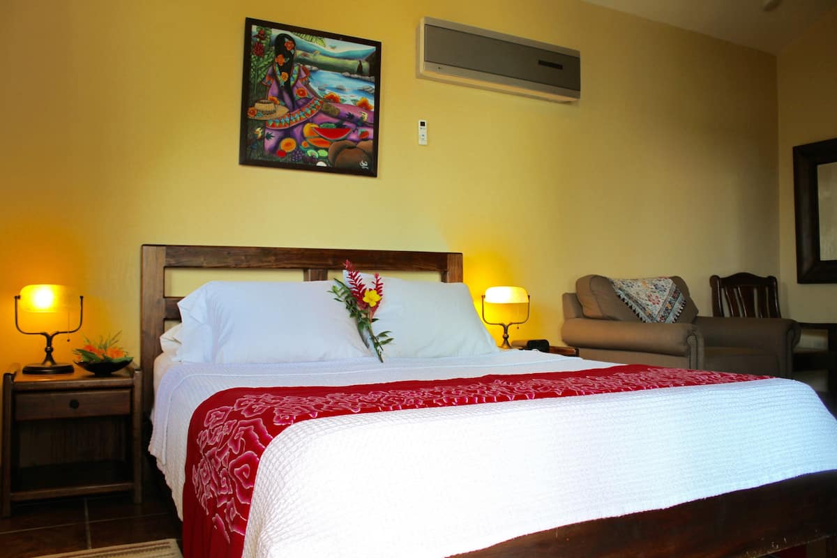 A neatly made bed with a red and white bedspread, a nightstand with a lamp, a chair, and a painting on the wall.