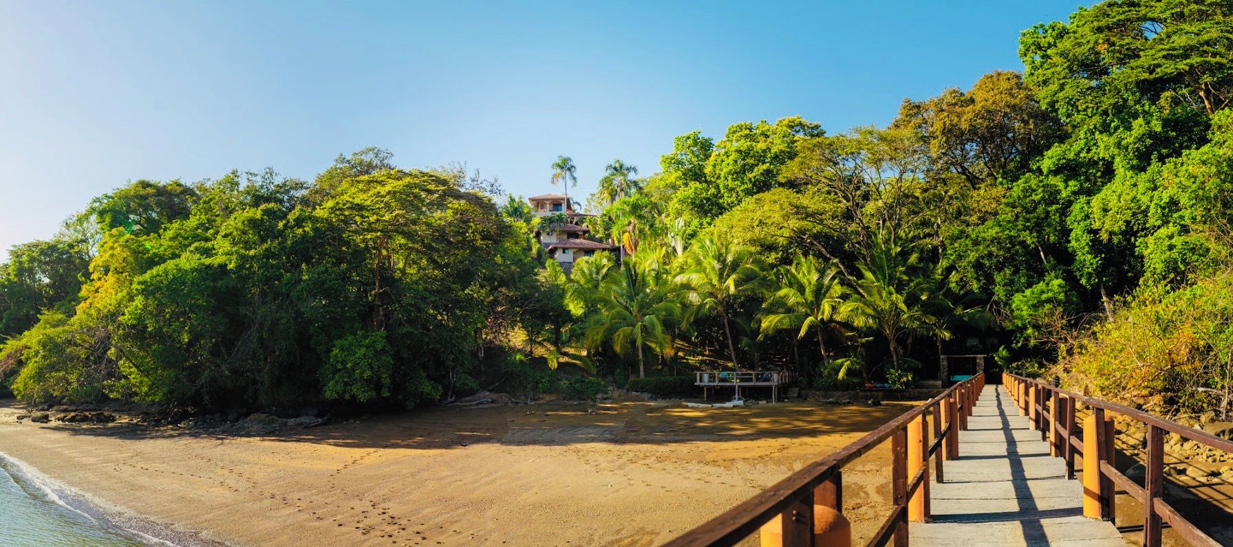 Une plage avec une promenade en bois menant à un bâtiment entouré d’une végétation luxuriante.