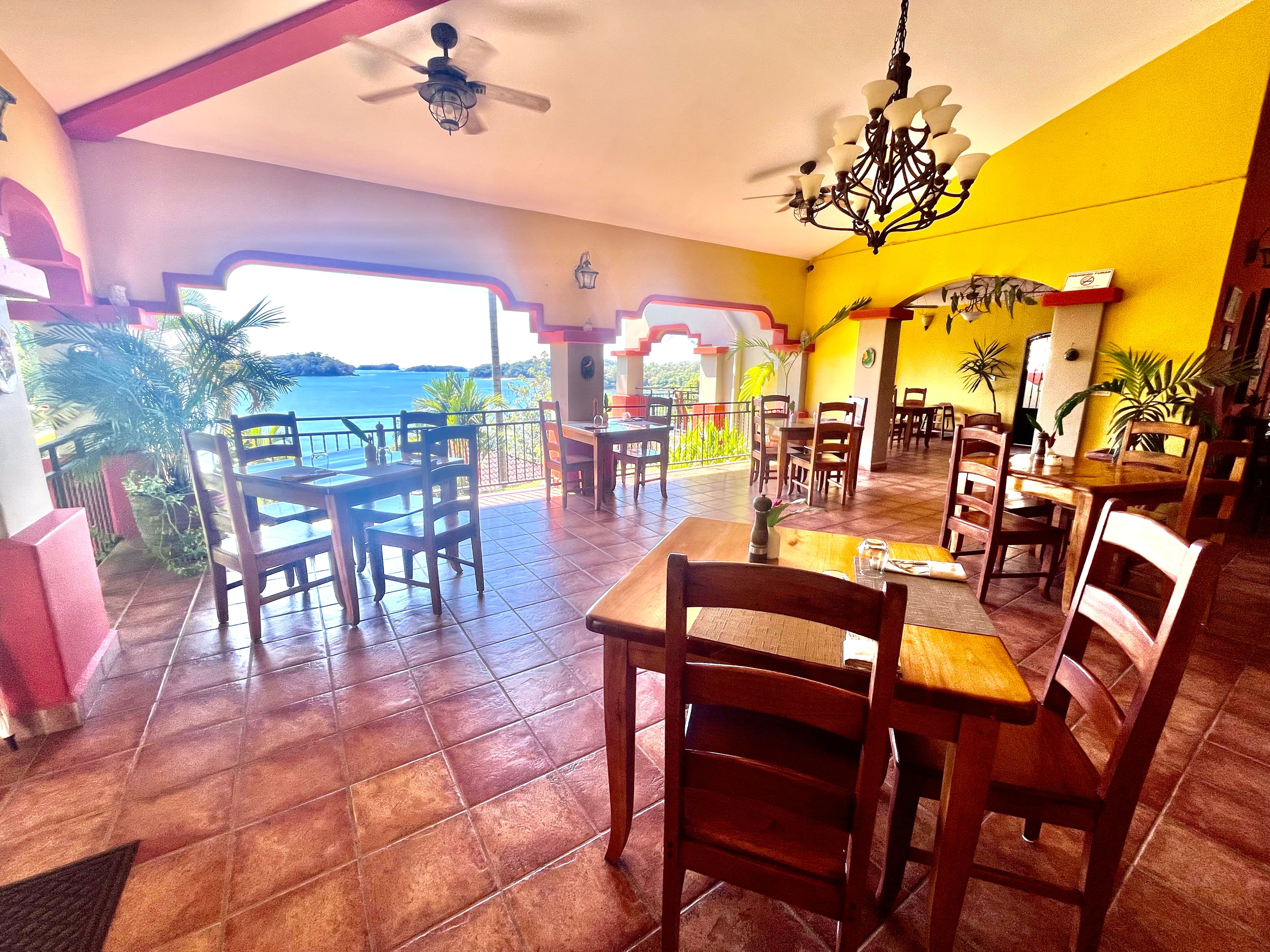 Restaurante com mesas e cadeiras de madeira, vista panorâmica, ventoinha no teto e lustre.