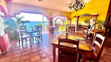 Restaurante com mesas e cadeiras de madeira, vista panorâmica, ventoinha no teto e lustre.