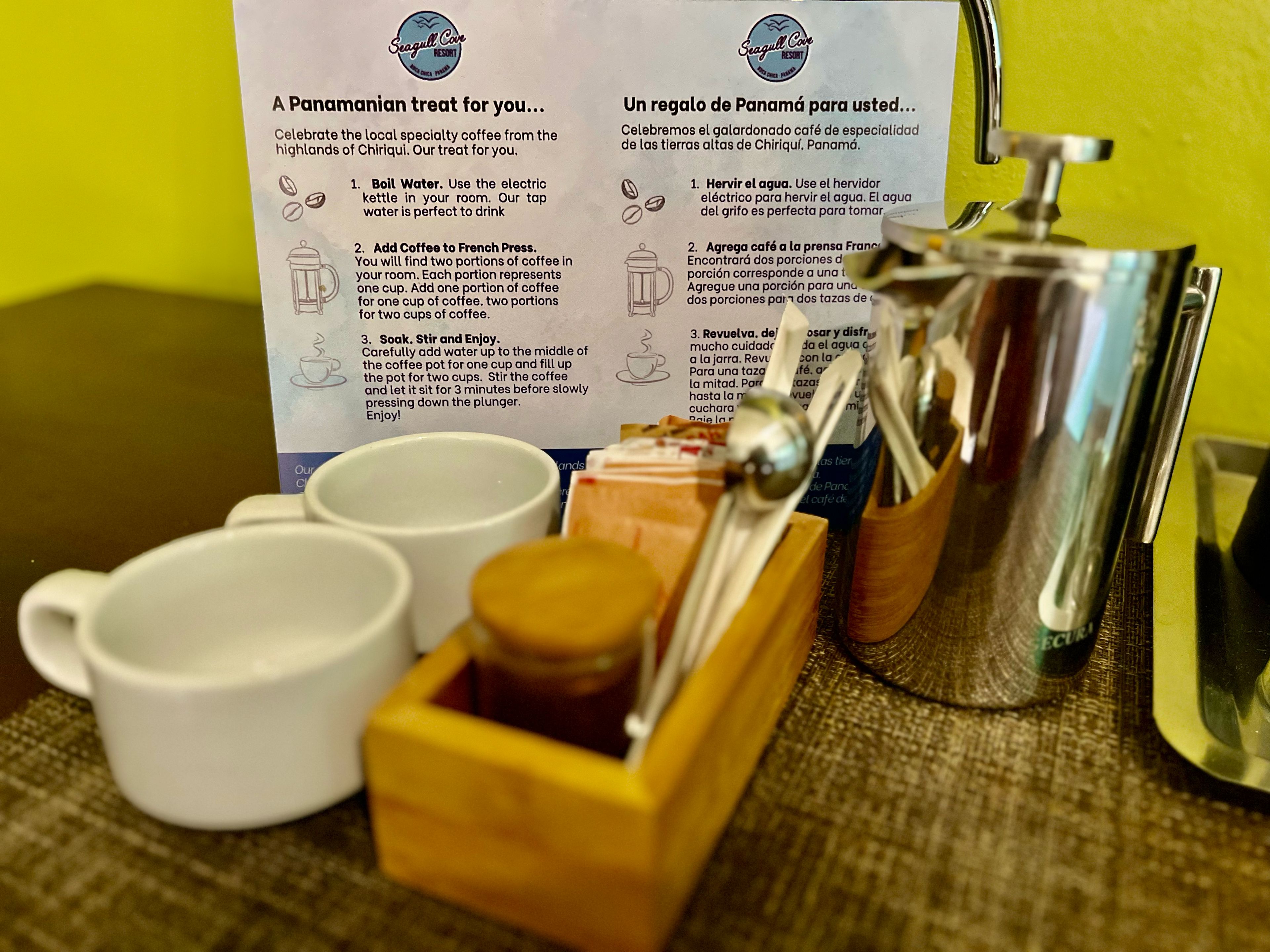 A coffee-making setup with a French press, two cups, and a wooden box with coffee beans. Instructions for making coffee are visible on a sign.