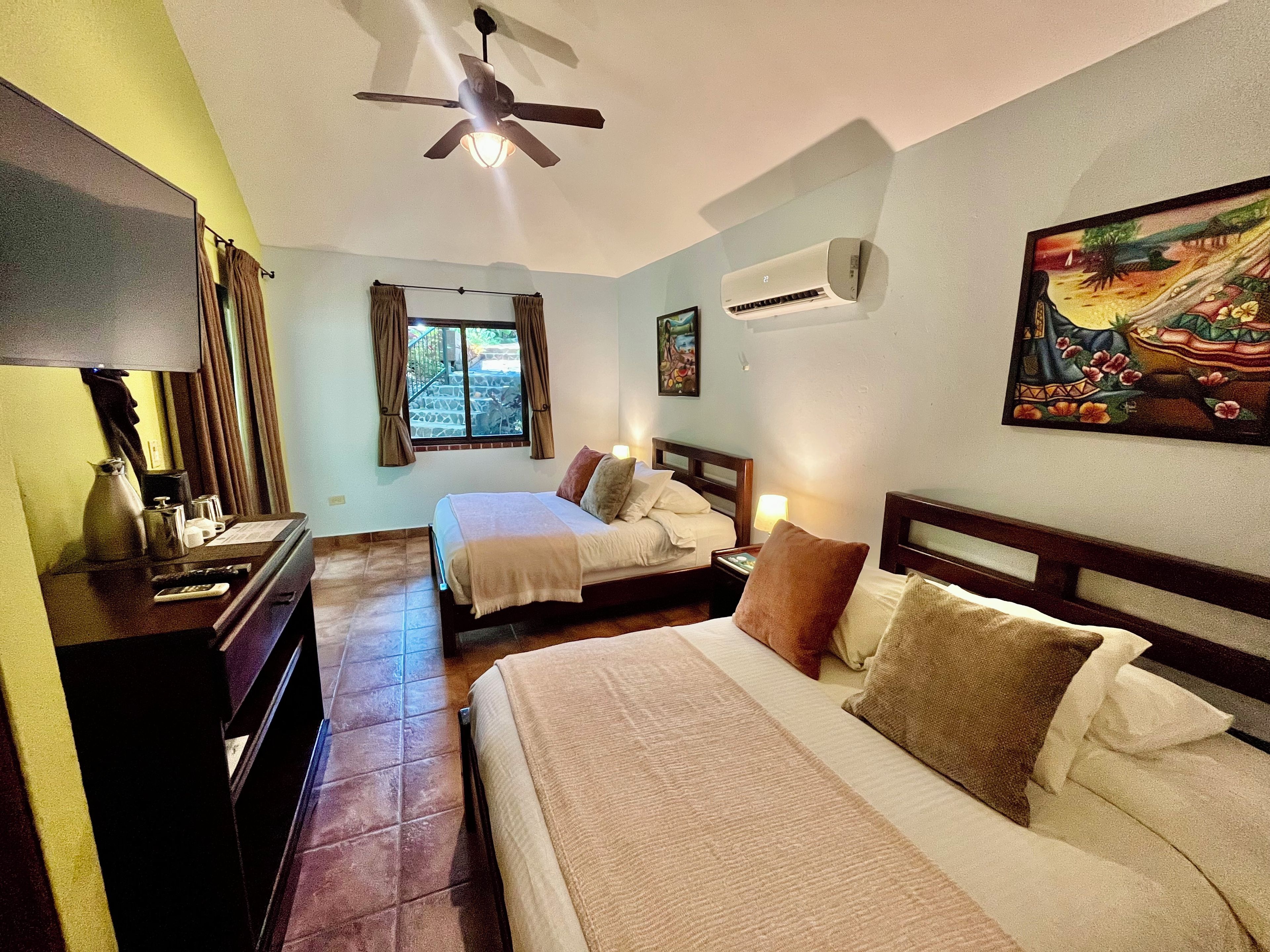 Une chambre d’hôtel avec deux lits, un ventilateur de plafond, un appareil de climatisation, un tableau au mur et une fenêtre avec des rideaux.