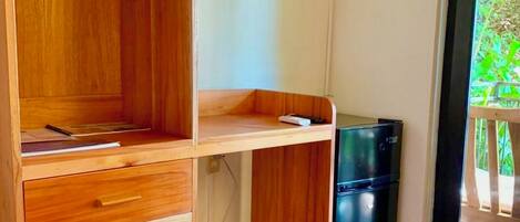Una habitación con un escritorio de madera, un televisor montado en la pared, un refrigerador pequeño y una puerta corrediza de vidrio que da a un área exterior.