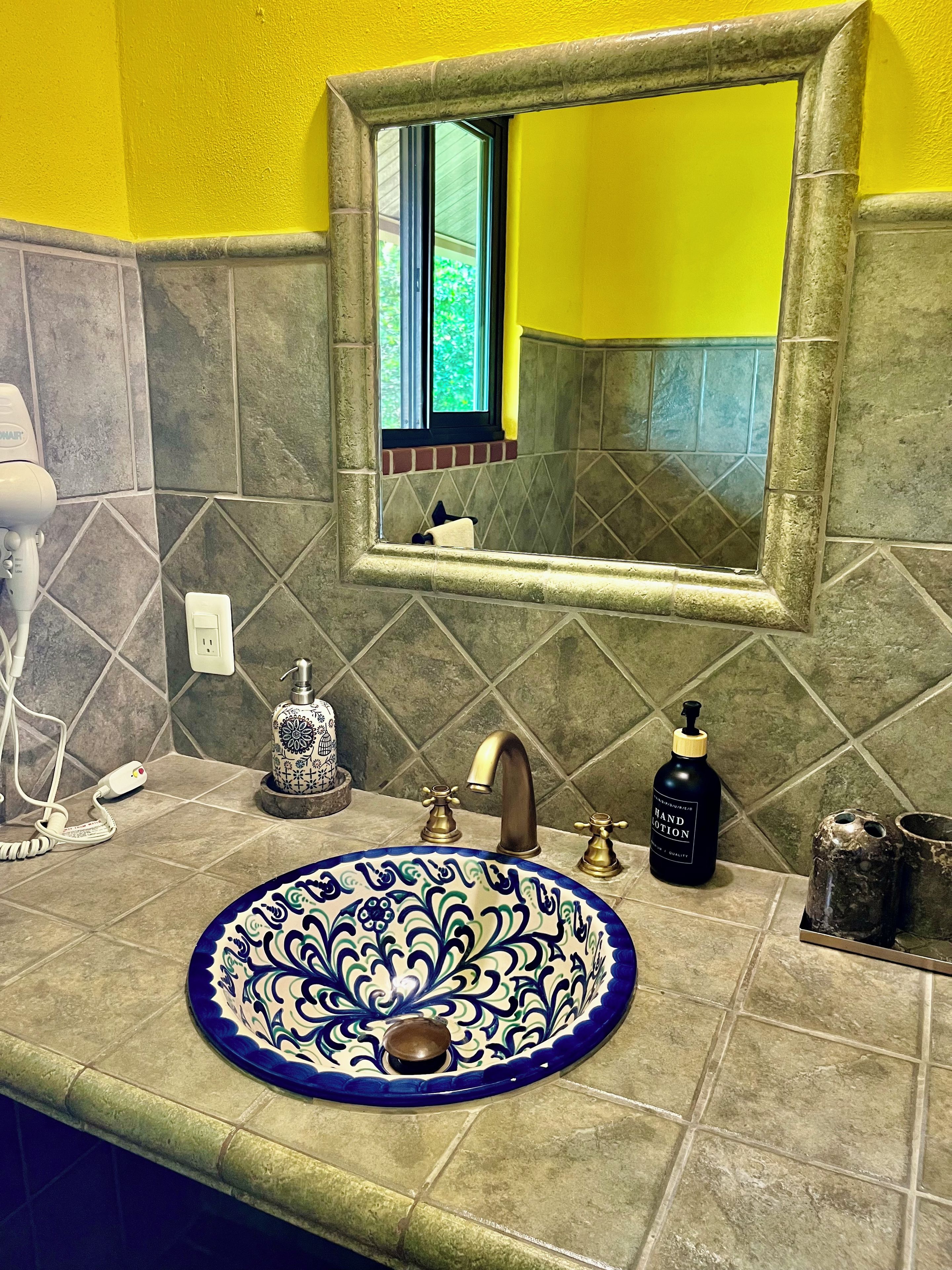 A bathroom with a yellow wall, a mirror, a sink with a blue and white patterned basin, a handheld hair dryer, a soap dispenser, and a bottle of hand lotion.