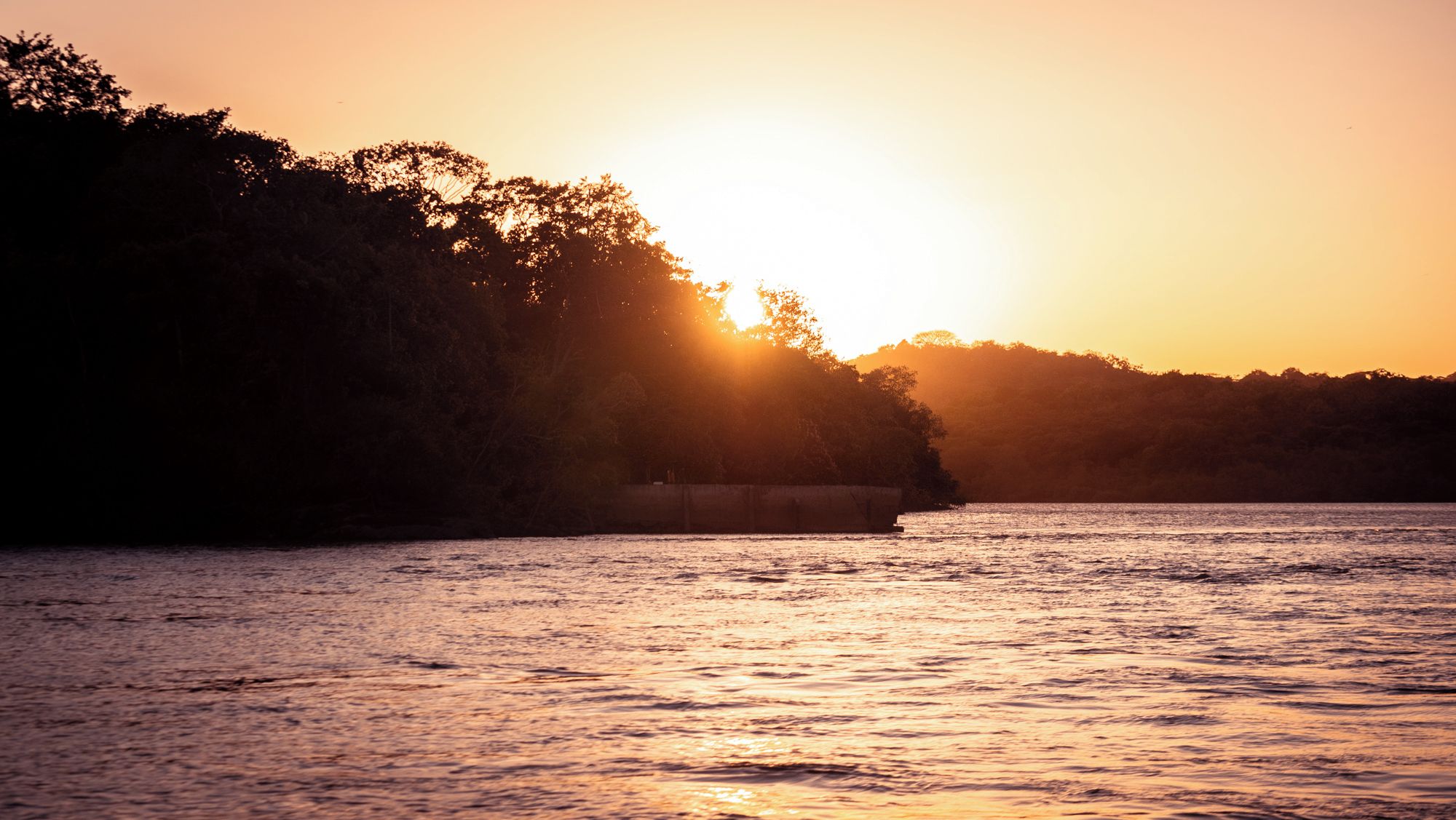 Atardecer sobre una masa de agua con la silueta de una tierra cubierta de árboles.