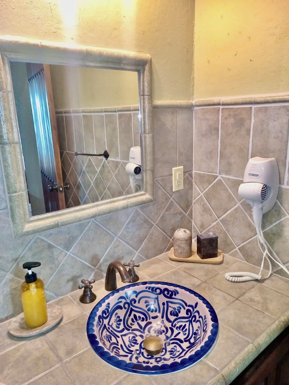 Un baño con espejo, un lavabo con lavamanos estampado en azul, un dispensador de jabón amarillo, un secador de pelo y un perchero para toallas.