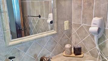 Un baño con espejo, un lavabo con lavamanos estampado en azul, un dispensador de jabón amarillo, un secador de pelo y un perchero para toallas.