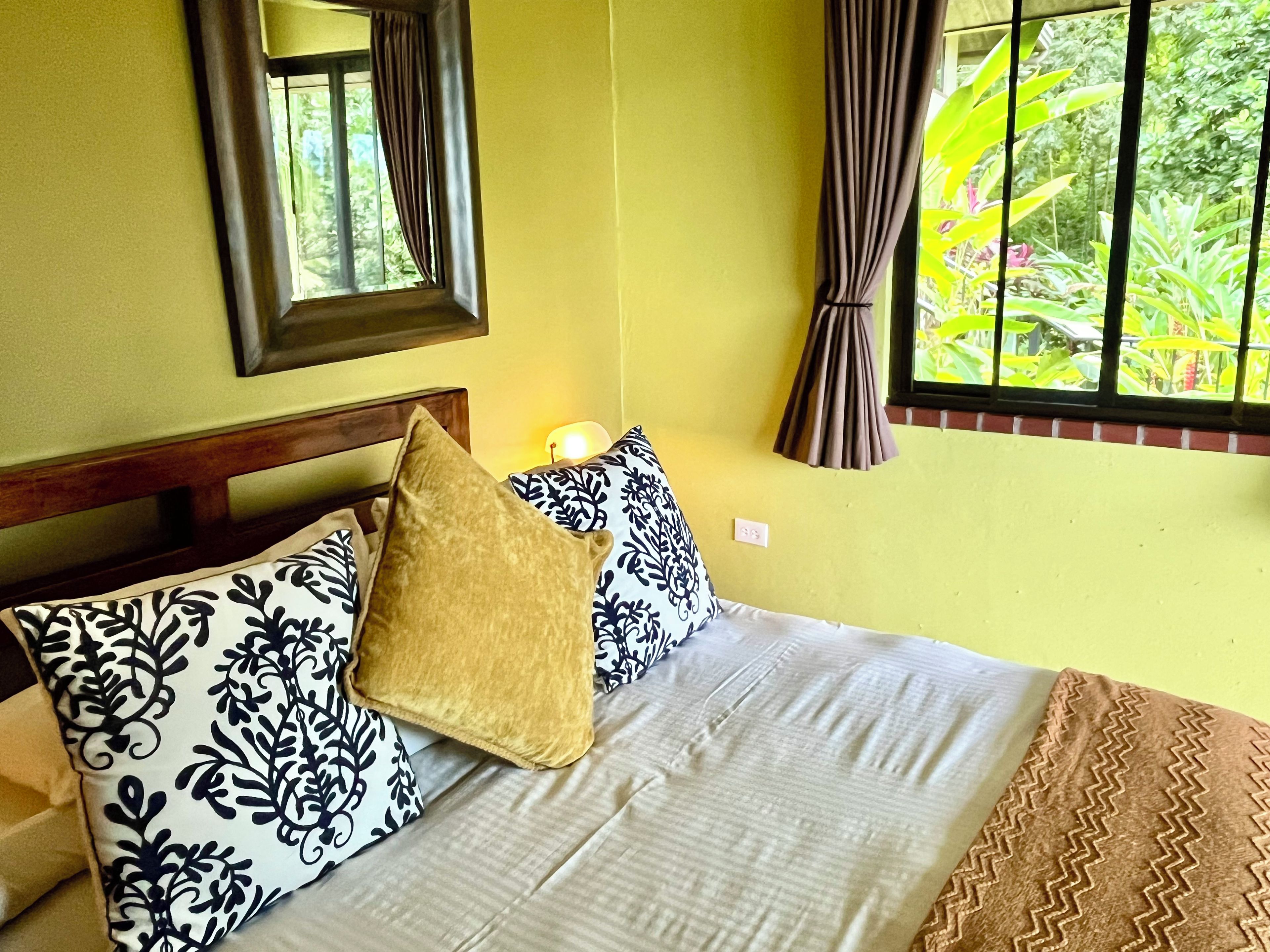 Una cama con almohadas estampadas, un cabecero de madera y una ventana con cortinas.
