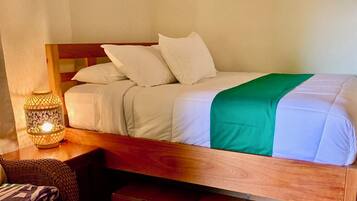 Un lit avec une literie blanche et un plaid vert, une table de chevet avec une lampe, et une chaise en osier.