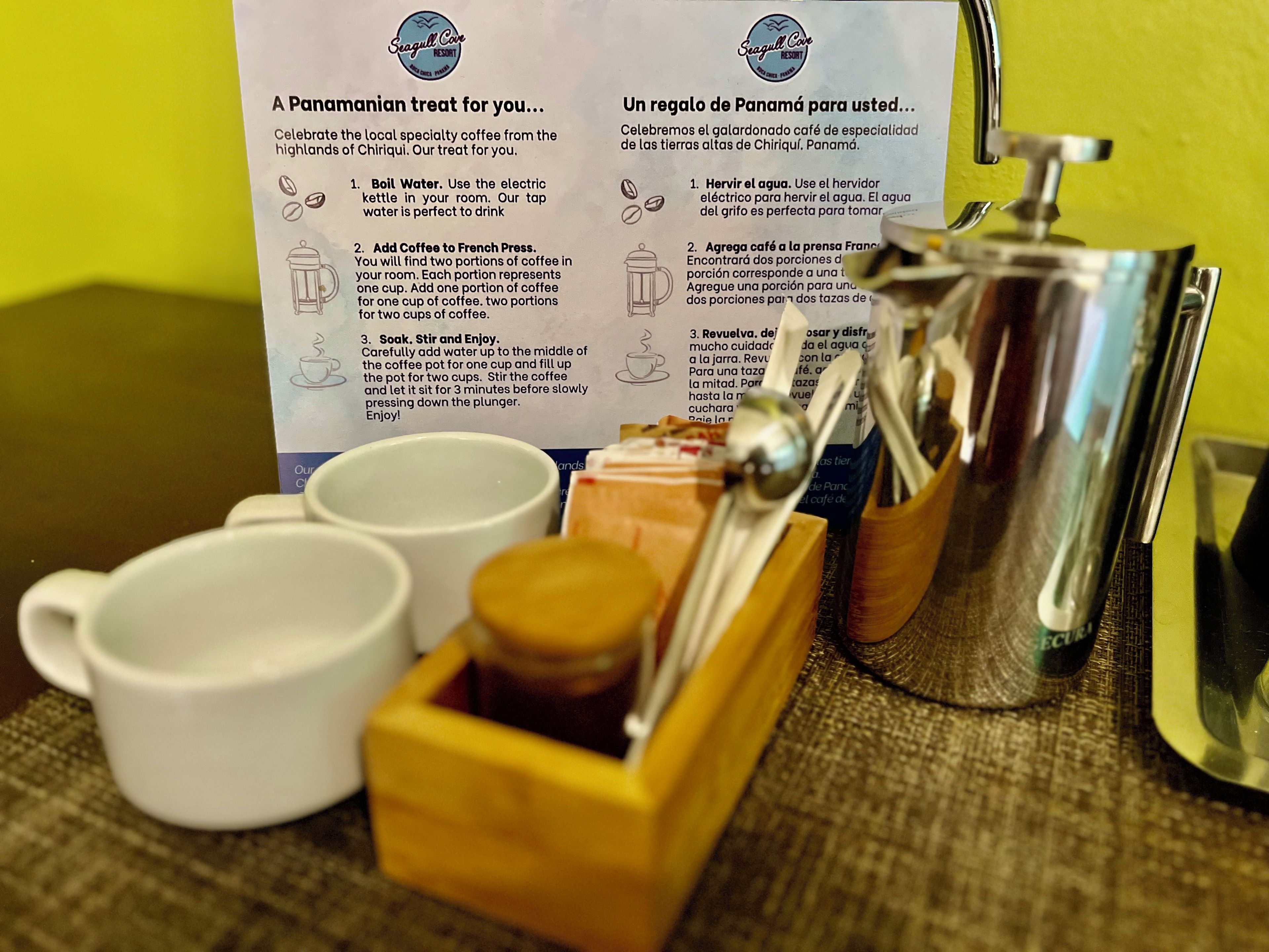 Een koffiezetapparaat met een French press, twee kopjes en een houten doos met koffiebonen. Op een bord staan instructies voor het zetten van koffie.