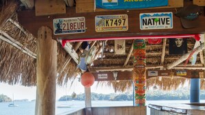 Una terraza de madera con taburetes de bar, techo de paja y vista al agua.