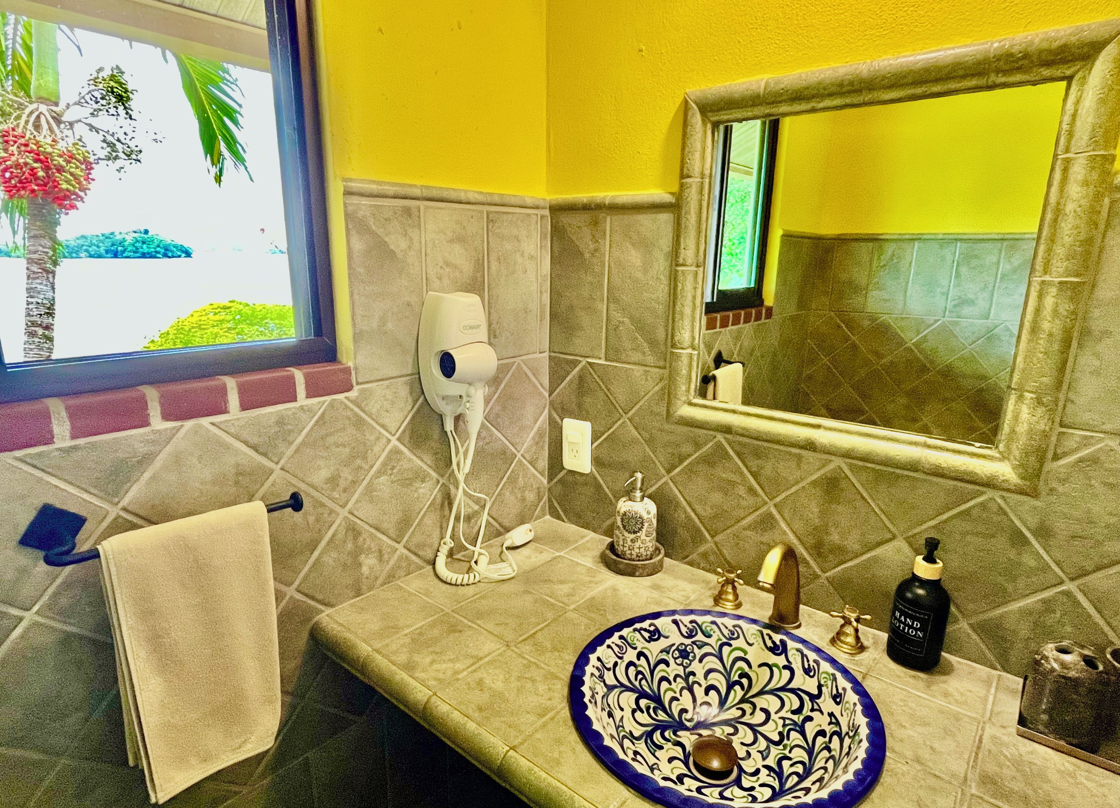 A bathroom with a sink, mirror, towel rack, and a window with a view of palm trees.