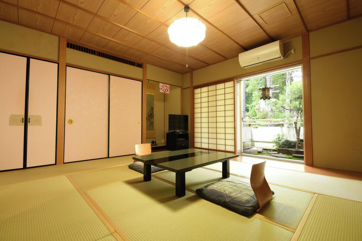 A traditional Japanese room with tatami flooring, a low table, a cushion, sliding doors, and a view of greenery outside.