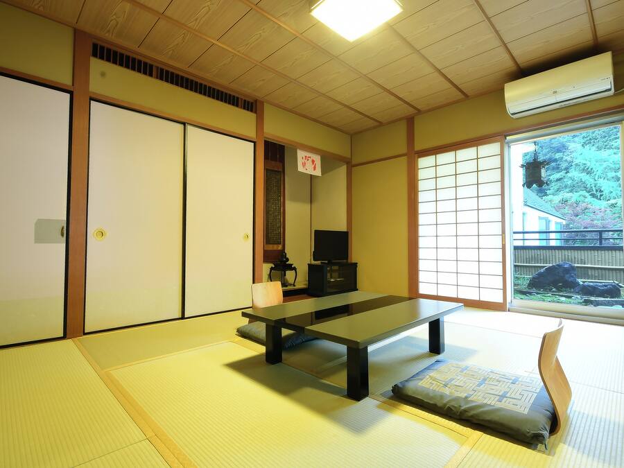 A traditional Japanese room with tatami flooring, a low table, sliding doors, and a view of a garden.