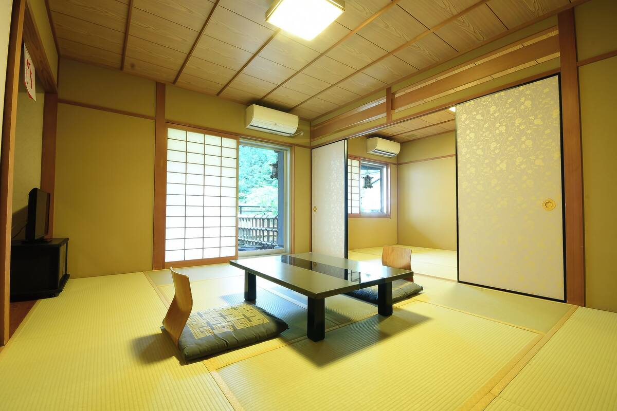A traditional Japanese room with tatami flooring, a low table, cushions, sliding doors, and a view of a garden.