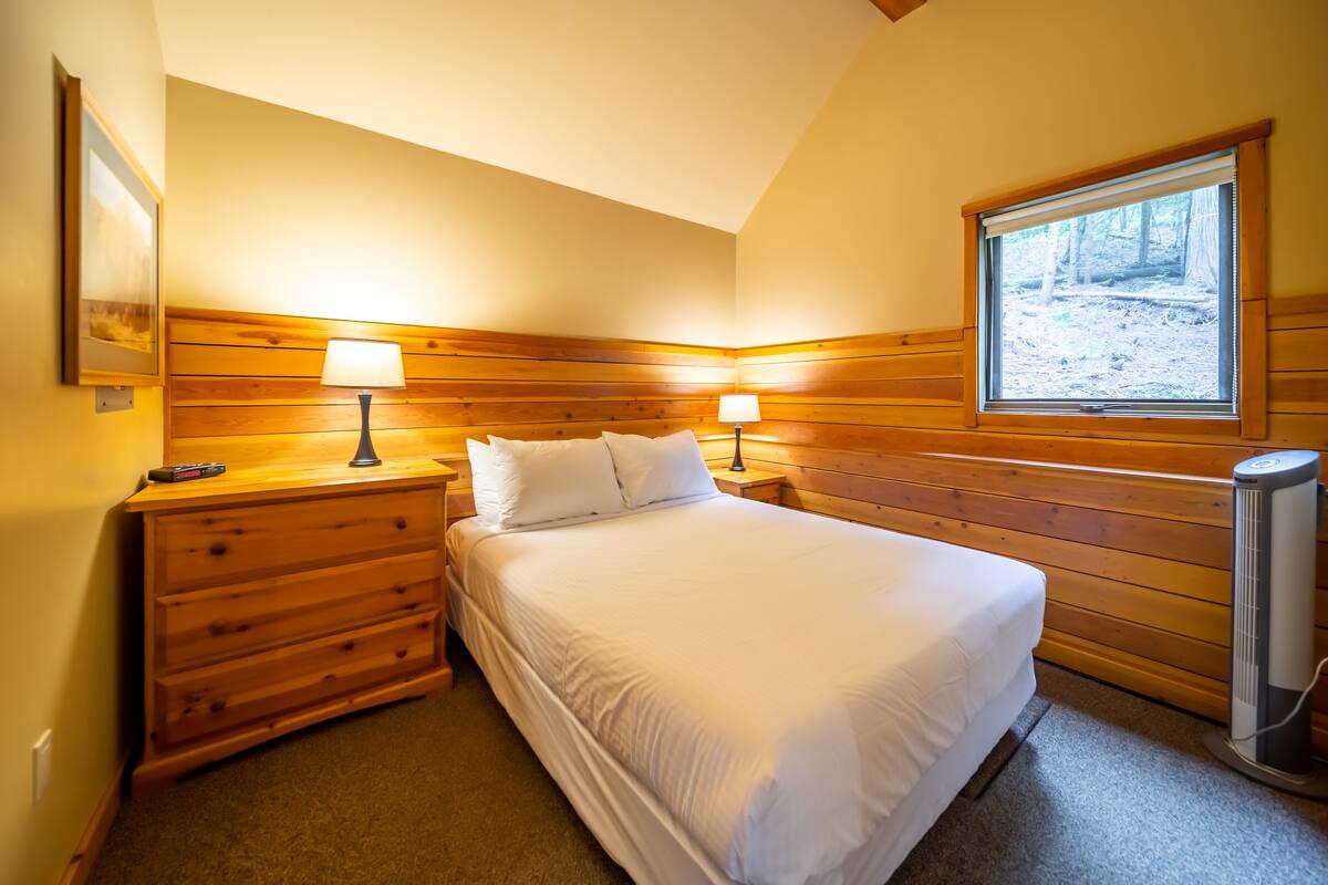 A bedroom with a wooden headboard, two bedside tables with lamps, a bed with white bedding, a window with a view of a forest, and a heater on the floor.