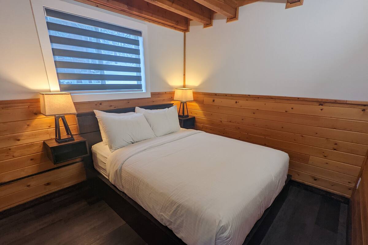 A bedroom with a wooden headboard, a bed with white bedding, two bedside lamps, and a window with blinds.