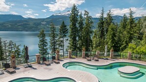 A swimming pool with a curved design, surrounded by lounge chairs and a stone deck, overlooking a lake and forested mountains. - Halcyon Hot Springs Resort (Nakusp)