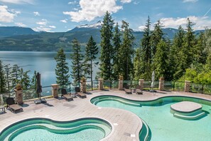 A swimming pool with a curved design, surrounded by lounge chairs and a stone deck, overlooking a lake and forested mountains. - Halcyon Hot Springs Resort (Nakusp)