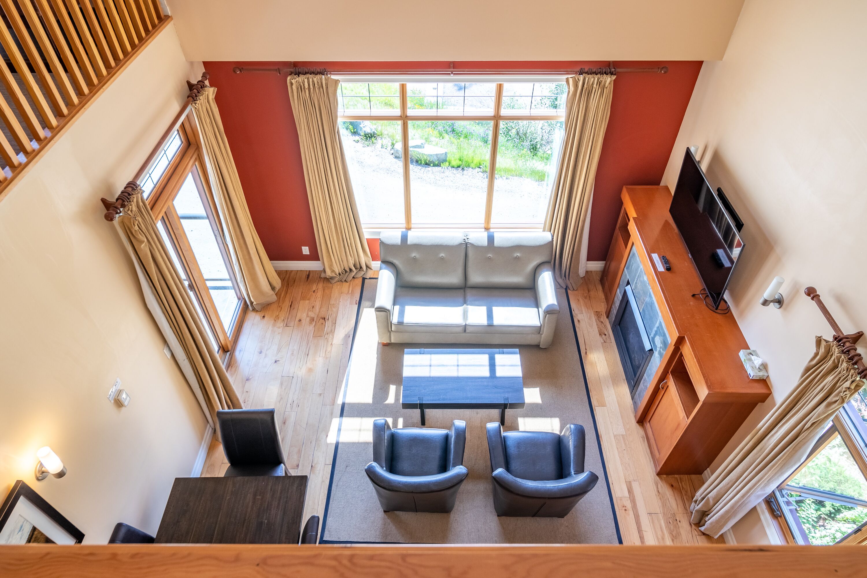 Una habitación con techos altos, un sofá, dos sillones, una mesa de centro y un televisor. Amplias ventanas con cortinas ofrecen vistas al exterior.