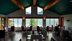 A dining area with tables set for guests, large windows offering views of a lake and mountains, and wooden beams on the ceiling. - Halcyon Hot Springs Resort (Nakusp)