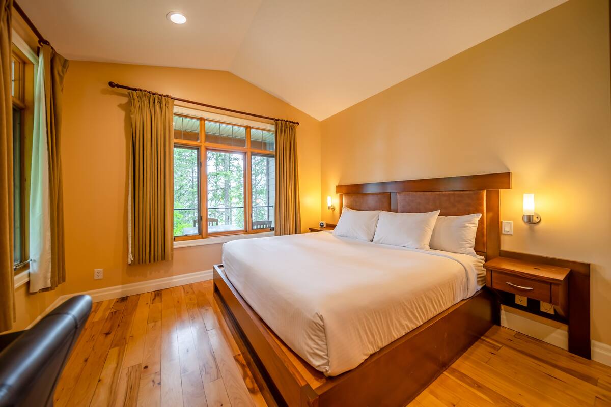 A large bed with a wooden headboard, a nightstand with a lamp, and a window with curtains.