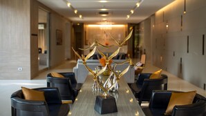 Uma zona de refeições moderna com uma mesa comprida, cadeiras pretas com almofadas douradas e uma escultura de metal decorativa no centro.