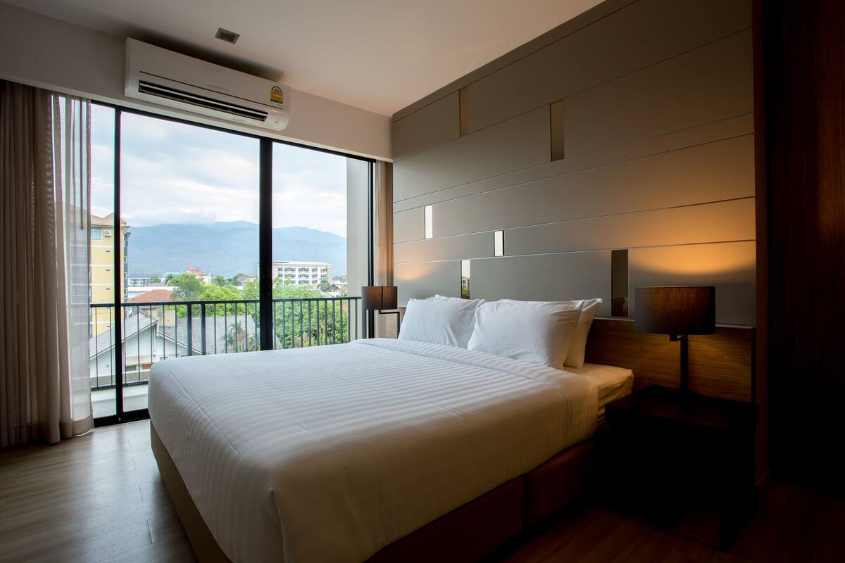 Habitación de hotel con una cama grande, una mesita de noche, una lámpara y vistas a la ciudad a través de amplias ventanas.