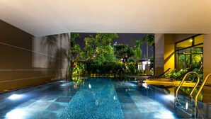 Uma piscina interior moderna com água cristalina, rodeada por vegetação exuberante e palmeiras, com vista para o céu noturno.